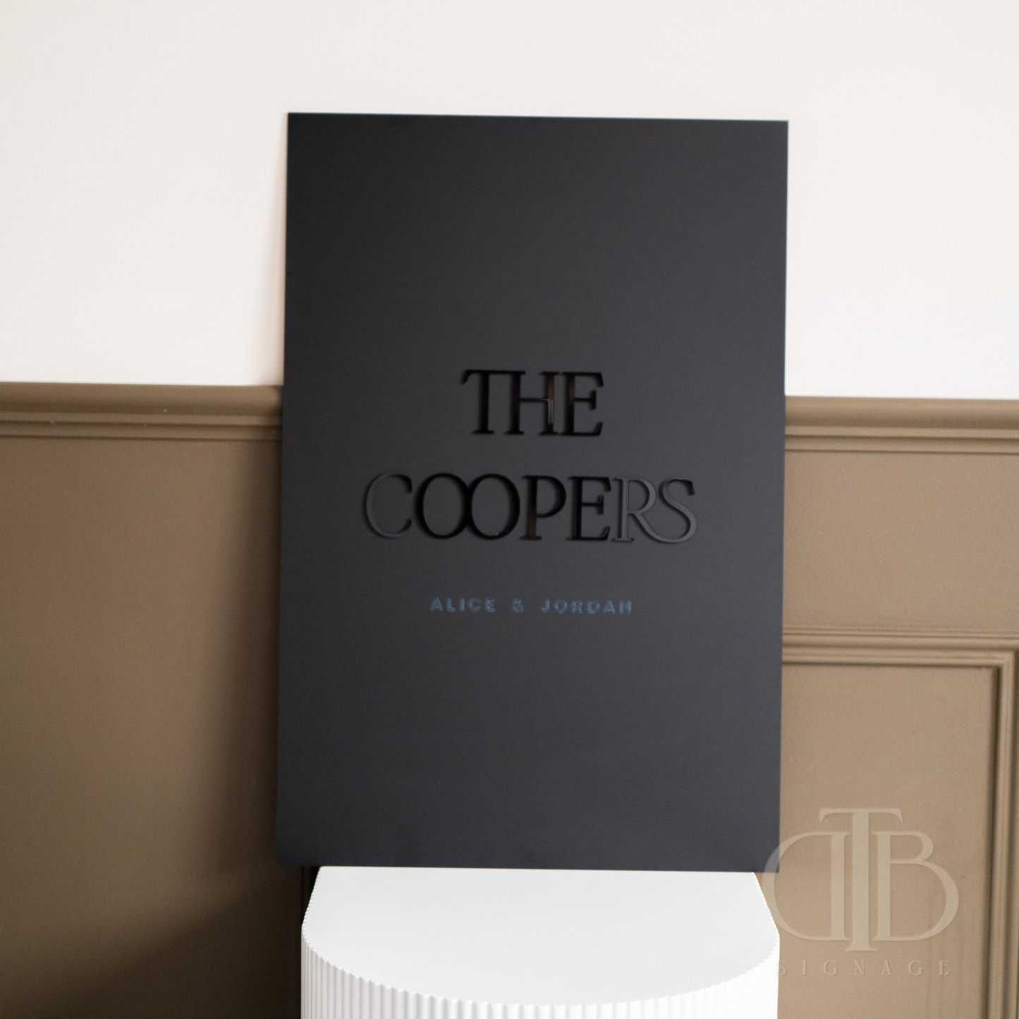 Black book titled 'The Coopers' by Alice & Jordan on a white pedestal against a beige wall.