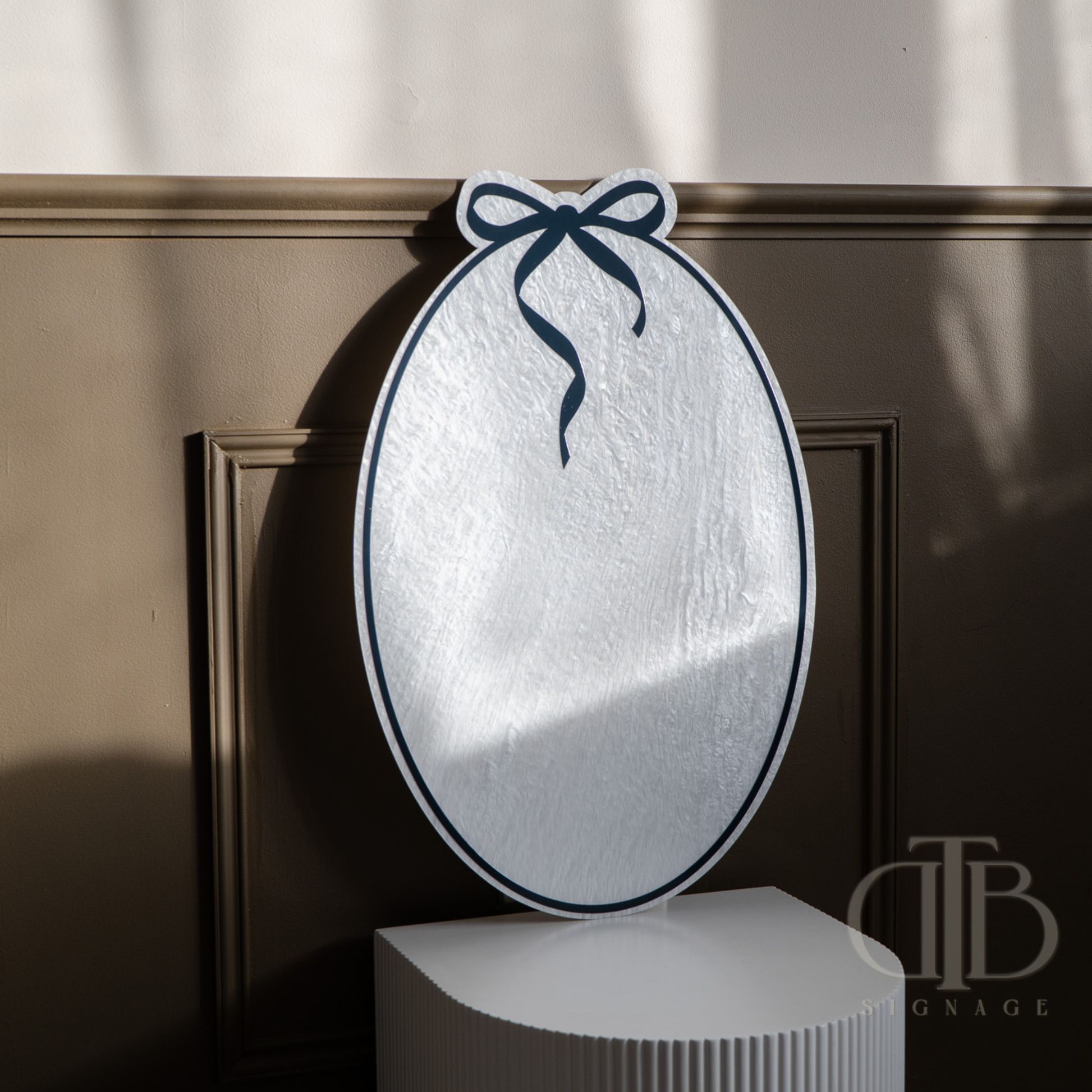 Decorative mirror with a bow on a wooden surface, featuring the brand 'DB Signage'.