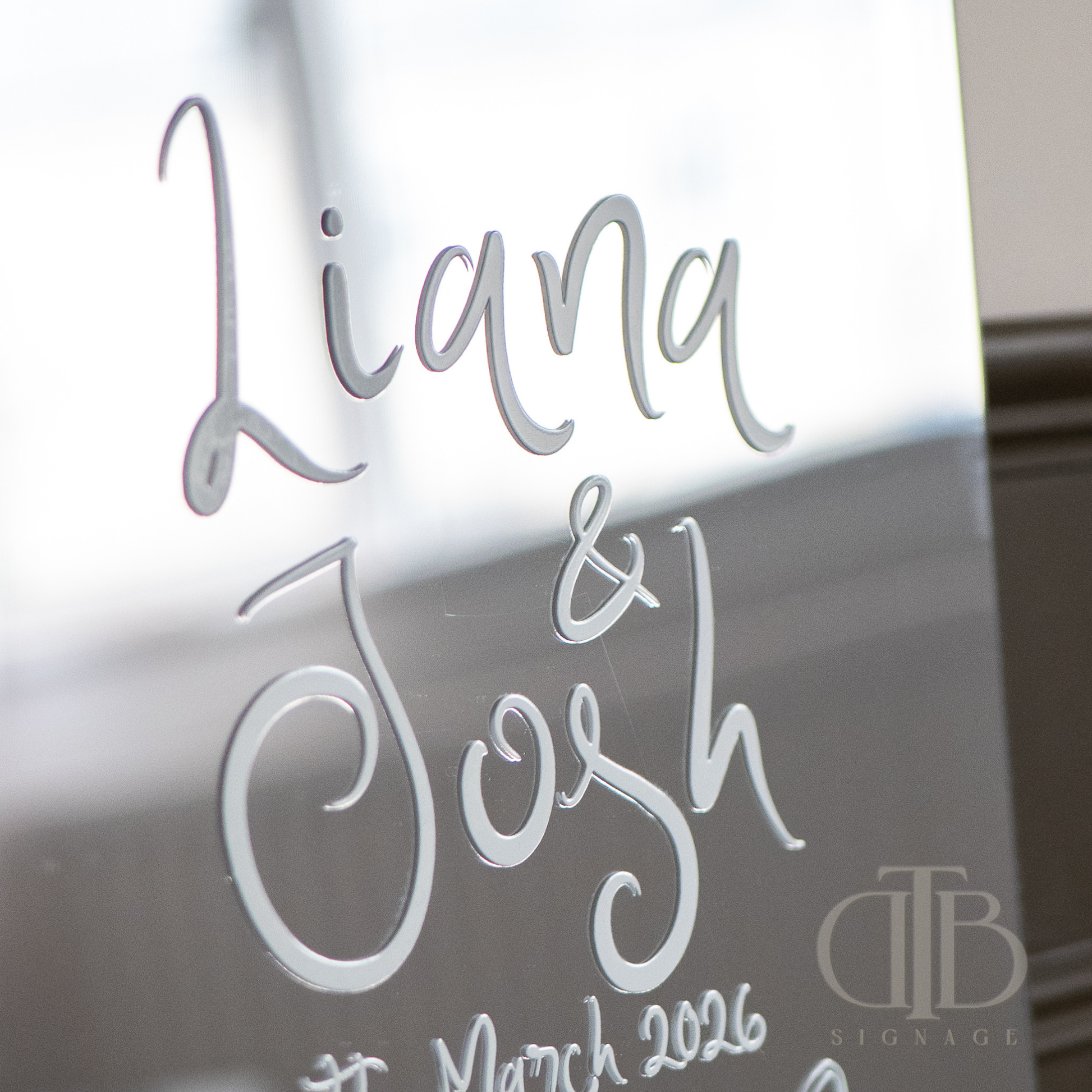 Decorative glass with engraved names 'Liana & Josh' and date 'March 2026' on a reflective surface.