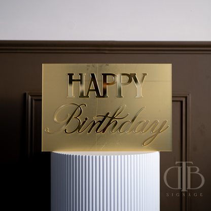 Gold 'Happy Birthday' sign on a white cylindrical object against a dark background