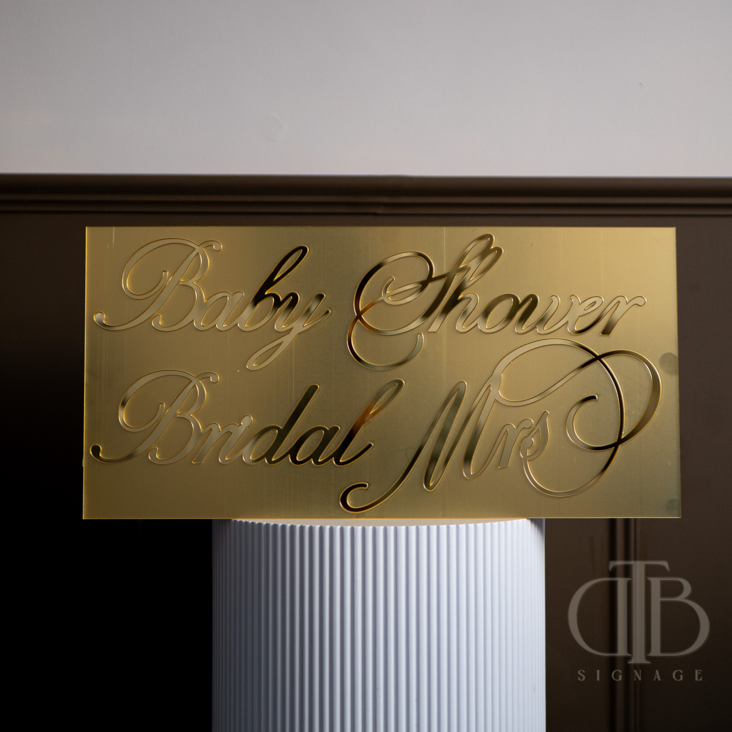 Gold sign with 'Baby Shower Bridal Mrs' text on a white pedestal