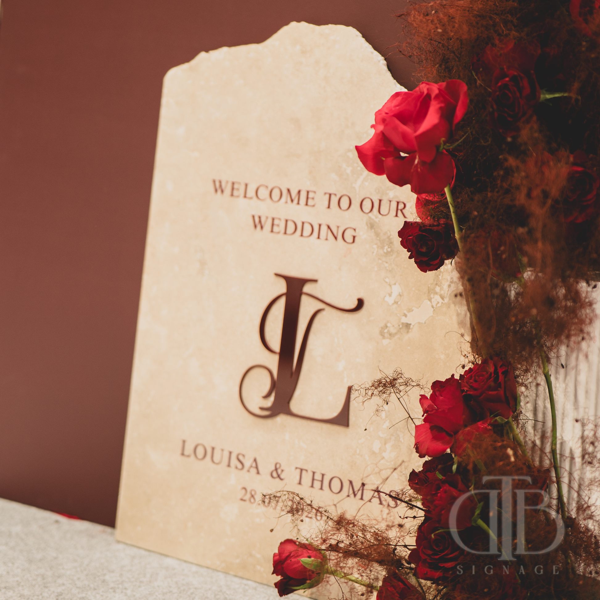Wedding welcome sign with red roses and 'L' monogram on a textured surface.