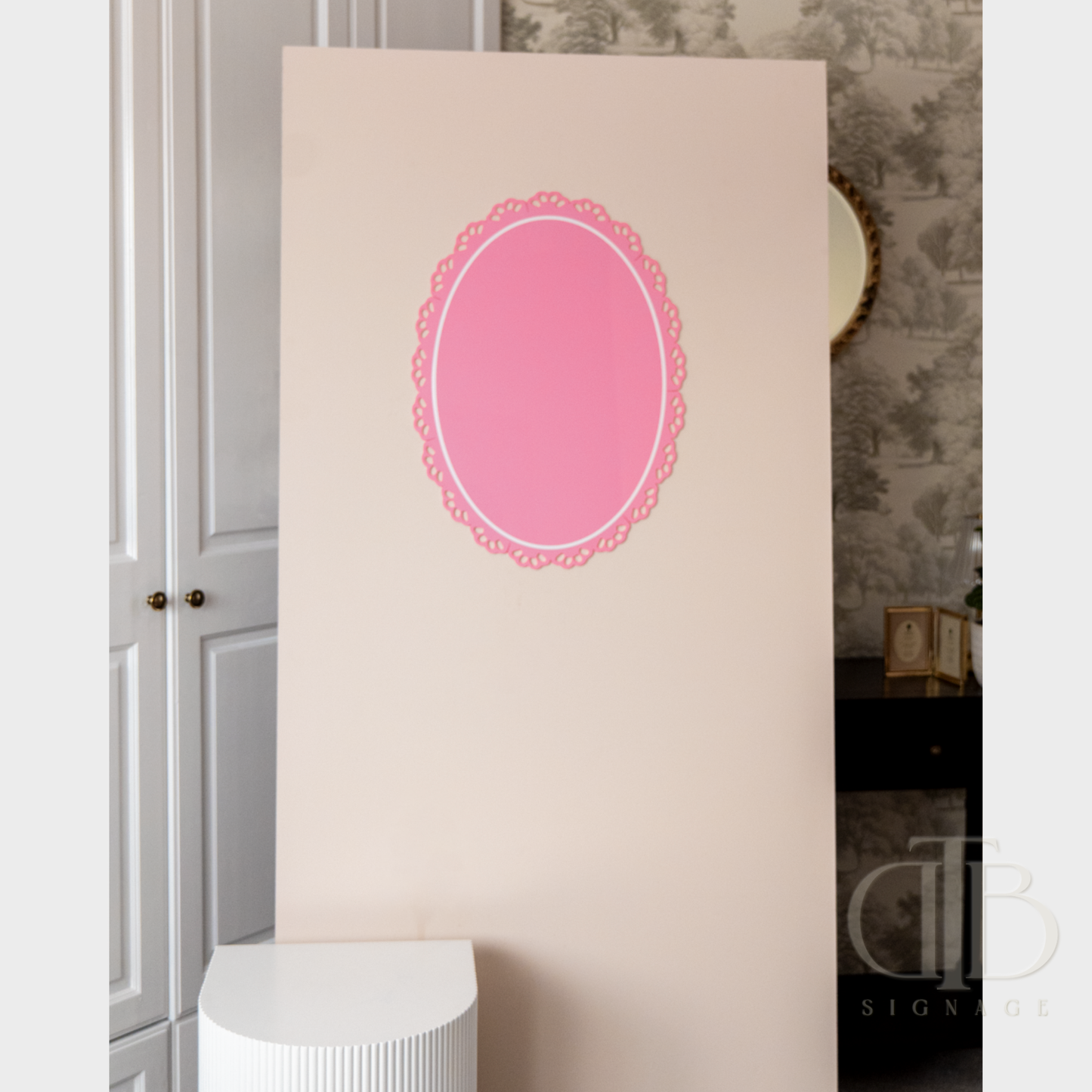 Pink oval acrylic signage on a beige wall with a textured gray wall and white cabinet in the background.