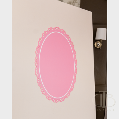 Pink oval acrylic signage with lace edge on a white surface