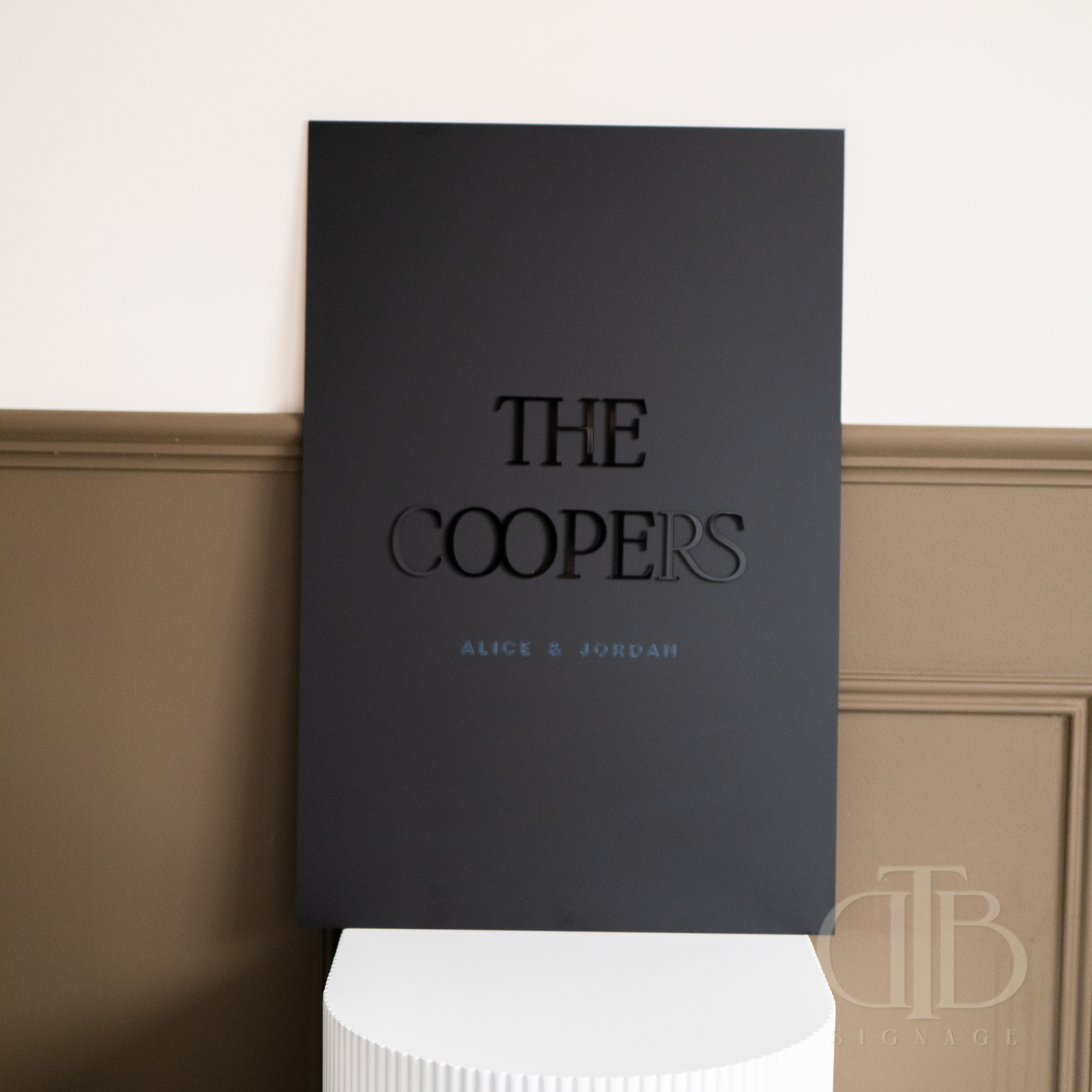 Black book titled 'The Coopers' by Alice & Jordan on a white pedestal against a beige wall.