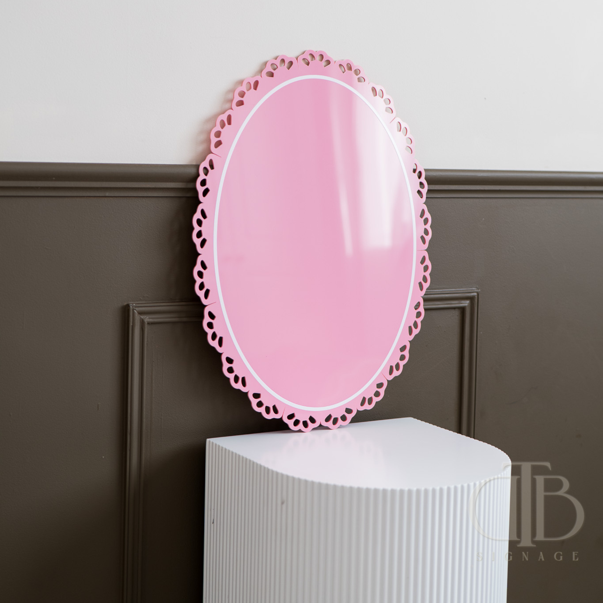 Pink oval acrylic signage with decorative frame on a white pedestal against a dark paneled wall.