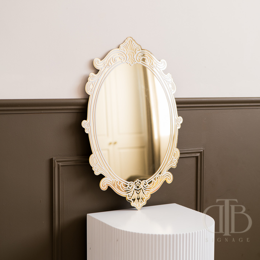 Decorative oval mirror with intricate frame on a white pedestal against a brown wall.