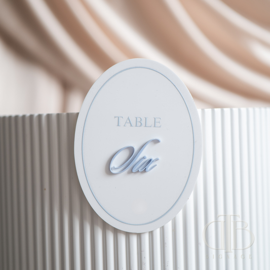 Oval acrylic table signage stuck on a white rippled plinth with beige drape background.