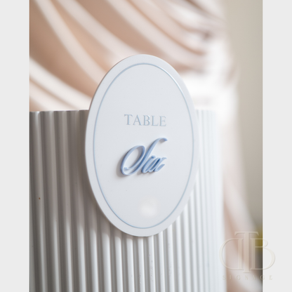 Oval acrylic table signage stuck on a white rippled plinth with beige drape background.